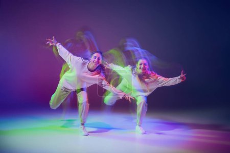 Portrait of young girls dancing hip-hop isolated over gradient blue purple background in neon with mixed light. Hobby. Concept of movement, youth culture, active lifestyle, action, street dance