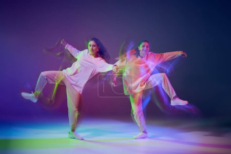 Foto de Retrato de chicas jóvenes bailando hip-hop aisladas sobre fondo azul púrpura degradado en neón con luz mixta. Entretenimiento. Concepto de movimiento, cultura juvenil, estilo de vida activo, acción, danza callejera - Imagen libre de derechos