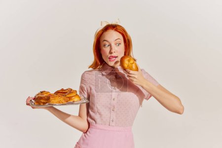 Téléchargez les photos : Portrait de jeune femme rousse en robe rose avec des petits pains fraîchement cuits isolés sur fond gris. Concept de beauté, style rétro, mode, élégance, années 60, 70, famille. Espace de copie pour la publicité - en image libre de droit