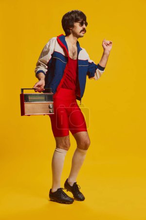 Un hombre con bigote posando en ropa deportiva vintage y pantalones cortos rojos aislados sobre fondo amarillo. Estilo de vida de los 90. Concepto de estilo retro, creatividad, emociones, expresión facial, moda