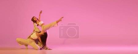 Two stylish female dancers in casual style clothes dancing contemporary choreography dance isolated over crystal pink background. Concept of modern art, creativity, fusion. Emotions in moves