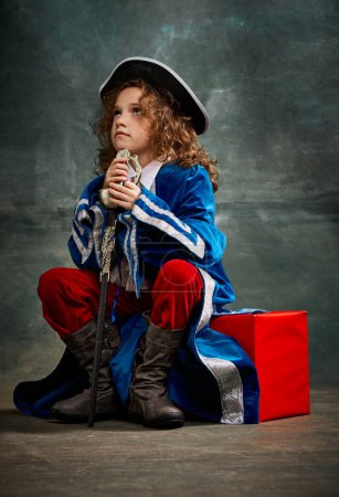 Photo for Thinking. Emotional kid, little girl wearing costume of prince, musketeer and royal person posing over dark vintage style background. Fashion, theater, beauty, emotions concept. Eras comparison - Royalty Free Image