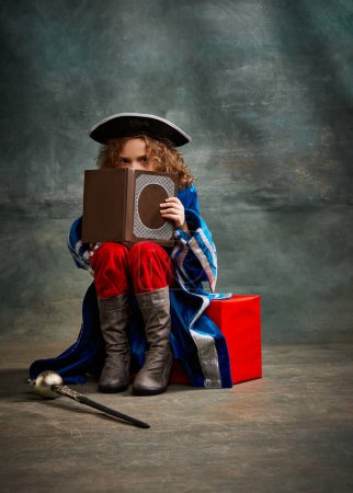 Photo for Kid reading book. Little girl wearing costume of prince, musketeer and royal person sitting and reading over dark vintage style background. Fashion, , education, emotions concept - Royalty Free Image
