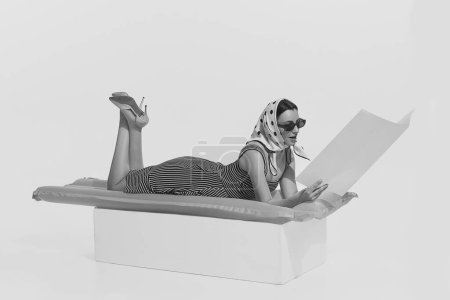 Foto de Vacaciones de verano. Retrato monocromático de una joven hermosa mujer acostada sobre un colchón inflable y una revista de lectura. Estilo vintage retro, moda de los años 70, 80. Copiar espacio para anuncio - Imagen libre de derechos