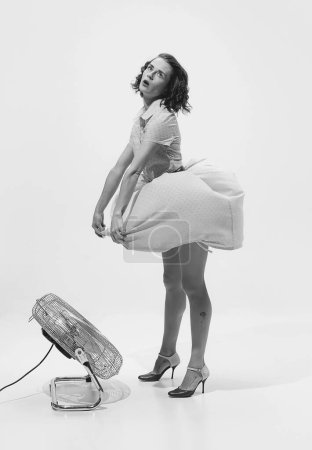 Téléchargez les photos : Portrait noir et blanc de jeune belle femme en robe rétro debout près du ventilateur. De chaudes journées d'été. Concept de beauté, style rétro, mode, élégance, années 60, 70, famille. Espace de copie pour la publicité - en image libre de droit