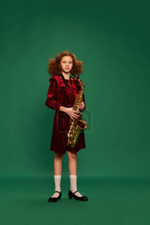 Téléchargez les photos : Bouclé rousse heureuse fille d'âge scolaire portant une robe de fête jouant sur le saxophone sur fond vert. Étudier, la mode, l'art, la musique et le style rétro créatif. Elle a l'air mignonne. - en image libre de droit