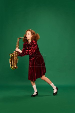 Téléchargez les photos : Curly redhaired happy school age girl wearing festive dress playing on saxophone over green background. Studying, fashion, art, music and creative retro style. Looks cute, funny - en image libre de droit