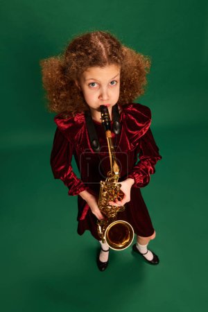 Téléchargez les photos : Wide angle view. Curly redhaired happy school age girl wearing festive dress playing on saxophone over green background. Studying, fashion, art, music and creative retro style concept - en image libre de droit
