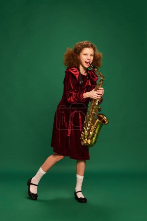 Téléchargez les photos : Curly redhaired happy school age girl wearing festive dress playing on saxophone over green background. Studying, fashion, art, music and creative retro style. Looks cute, funny - en image libre de droit