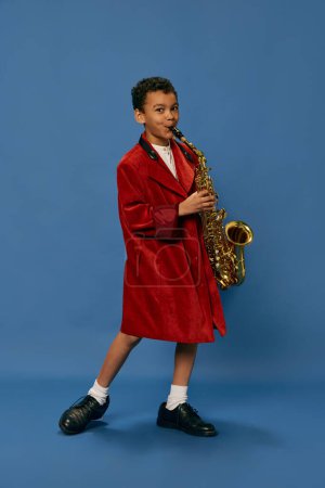 Téléchargez les photos : Like jazz man. Portrait of cute little african boy wearing huge mans jacket and shoes playing on saxophone over blue background. Fashion, art, music and creative style. Looks happy, cheerful - en image libre de droit
