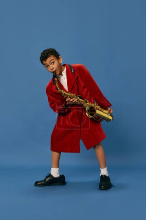 Téléchargez les photos : Comme un homme de jazz. Portrait de mignon petit garçon africain portant une énorme veste mans et des chaussures jouant sur le saxophone sur fond bleu. Mode, art, musique et style créatif. Ça a l'air heureux, joyeux - en image libre de droit
