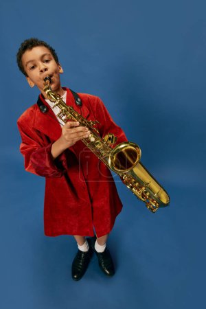 Téléchargez les photos : Like jazz man. Wide angle view image of cute little african boy wearing huge mans jacket and shoes playing on saxophone over blue background. Fashion, art, music and creative style. Melody - en image libre de droit