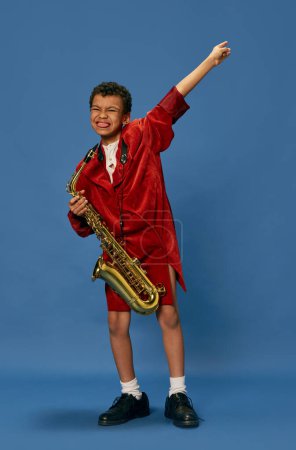Téléchargez les photos : Like jazz man. Portrait of cute little african boy wearing huge mans jacket and shoes playing on saxophone over blue background. Fashion, art, music and creative style. Looks happy, cheerful - en image libre de droit
