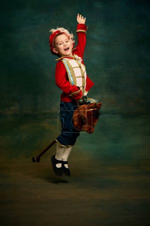 Photo for Happy cute little boy dressed up as medieval little prince and pageboy ride toy horse over dark vintage style background. Vintage fashion, emotions, theater art concept. Eras comparison - Royalty Free Image
