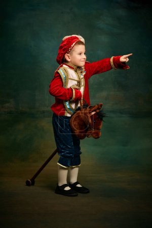 Photo for One cute little boy dressed up as medieval little prince and pageboy ride toy horse over dark vintage style background. Vintage fashion, emotions, theater art concept. Eras comparison - Royalty Free Image