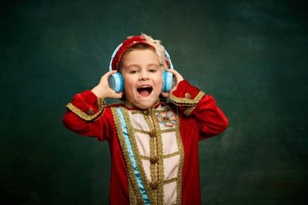 Photo for Cheerful little boy dressed up as medieval little prince and pageboy listening to music over dark vintage style background. Retro fashion, emotions, music concept. Kids looks happy - Royalty Free Image