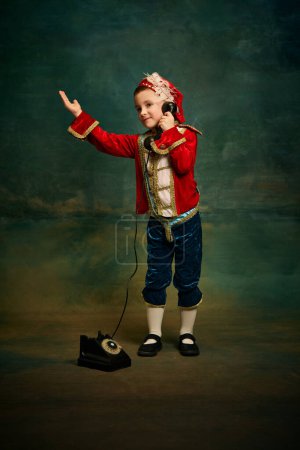 Photo for Charming little kid dressed up as medieval little prince and pageboy talking on old-fashioned phone over dark vintage style background. Eras comparison concept - Royalty Free Image