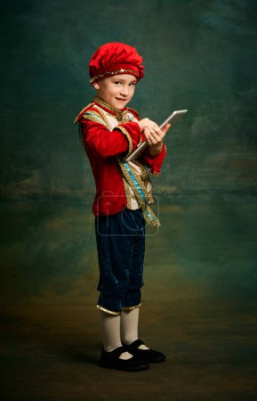 Photo for One cute little boy dressed up as medieval little prince and pageboy using tablet over dark vintage style background. Vintage fashion, emotions, theater art concept. Eras comparison - Royalty Free Image