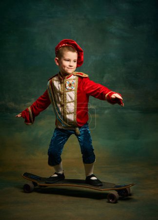 Photo for Skateboarder. Smiling little boy dressed up as medieval little prince and pageboy skateboarding over dark vintage style background. Action, leisure activities, emotions, sport concept - Royalty Free Image