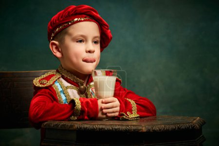 Photo for Tasting milk cocktail. Little charming kid in historical costume drinking milkshake over dark green background. Concept of fast food, comparison of eras, retro, vintage, emotions. Looks happy - Royalty Free Image