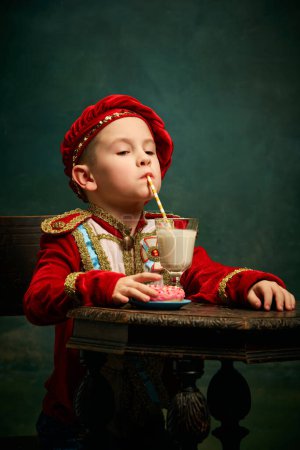 Photo for Tasting milk cocktail. Little charming kid in historical costume drinking milkshake over dark green background. Concept of fast food, comparison of eras, retro, vintage, emotions. Looks happy - Royalty Free Image