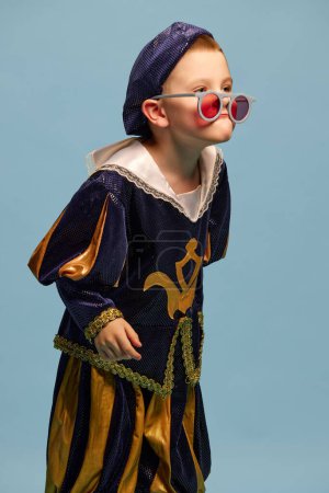 Photo for Portrait of little charming boy in costume of medieval pageboy, little prince having fun over light blue background. Concept of children emotions, eras comparison, fashion, theater, art, festival - Royalty Free Image