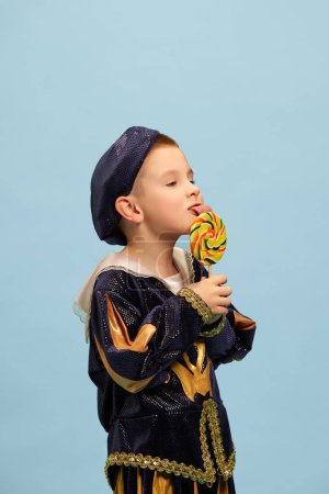 Photo for Sweets. Cute little charming boy in costume of medieval pageboy, little prince with big lollipop over light blue background. Concept of children emotions, eras comparison, fashion, theater - Royalty Free Image