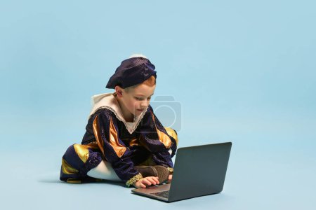 Photo for Online studying. Little cute boy wearing costume of medieval pageboy, little prince sitting in front of laptop over light blue background. Concept of children emotions, education, technology, ad - Royalty Free Image
