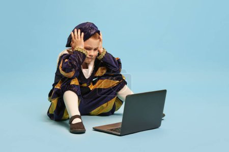 Photo for Bad news. Little cute boy wearing costume of medieval pageboy, little prince sitting in front of laptop over light blue background. Concept of children emotions, education, technology, ad - Royalty Free Image