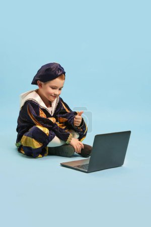 Photo for Chat with friends. Little cute boy wearing costume of medieval pageboy, little prince sitting in front of laptop over light blue background. Concept of children emotions, education, technology, ad - Royalty Free Image