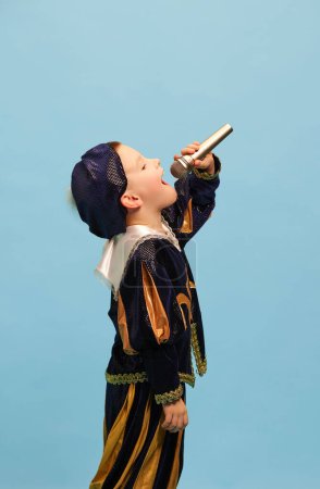 Photo for Happy preschool age boy in costume of medieval pageboy, little prince singing at microphone over light blue background. Concept of children emotions, eras comparison, fashion, music, art, festival - Royalty Free Image
