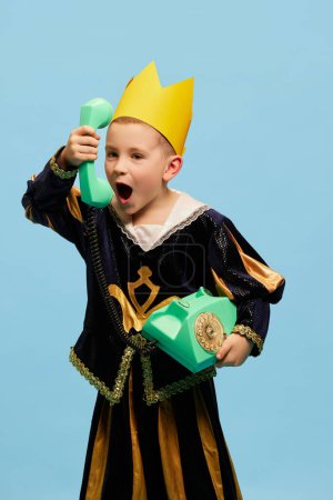 Photo for Joyful little cute boy in costume of medieval little prince in paper crown talking on retro phone over light blue background. Concept of children emotions, eras comparison, vintage fashion, ad - Royalty Free Image