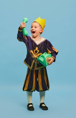 Photo for Joyful little cute boy in costume of medieval little prince in paper crown talking on retro phone over light blue background. Concept of children emotions, eras comparison, vintage fashion, ad - Royalty Free Image