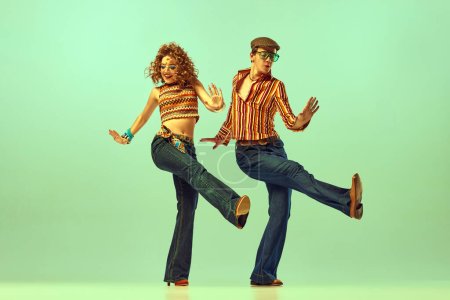 Dynamisches Porträt glücklicher Männer und Frauen in Retro-Hemden und ausgestellten Jeans, die energiegeladenen Tanz über grünem Hintergrund tanzen. Konzept der Modetrends der 70er, 80er Jahre, Musik, Hippie-Lifestyle