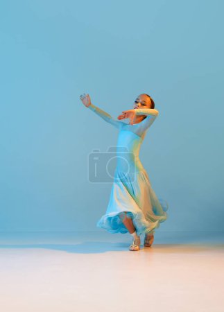 Waltz. Portrait of little flexible girl in stage outfit, dress dancing ballroom dance over blue background. Concept of beauty, professional dances, skills. Emotions in movements