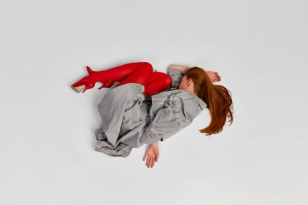 Impersonal human emotions. Stylish girl in grey coat and bright red tights moves over light background. Expressive fashion. Concept of art photography, beauty. Redhaired model in contemporary dance