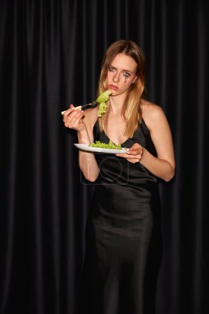 Photo for Portrait of misty and dramatic girl holding plate with lettuce and looking at camera with sad facial expression over black background. Concept of diet, beauty, healthy eating and harm - Royalty Free Image