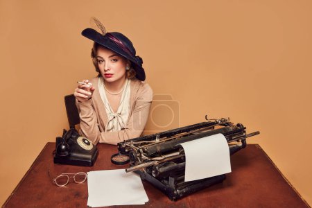 Photo for Calmness. Attractive woman writer wearing old-fashioned clothes sitting at table and smoking over beige background. Concept poems, novel, emotions, beauty, fashion, retro style, vintage - Royalty Free Image