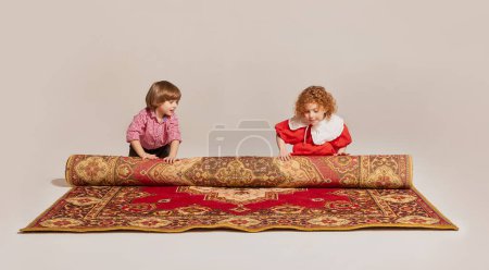 Photo for Excited kids. Portrait of charming little siblings wearing retro clothes and rolling up the carpet. Concept of happy childhood, leisure activities, fun, lifestyle, family, retro style, 60s, 70s - Royalty Free Image
