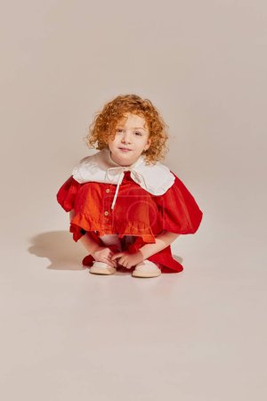 Photo for Charming. Cute little girl with curly redhead hair wearing dress and looking at camera over light background. Concept of carefree childhood, holidays, fun, human emotions, facial expression - Royalty Free Image