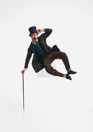 Levitating man. Portrait of a gentleman wearing old clothes and black cylinder jumping with cane up over white studio background. Concept of historical remake, comparison of eras, vintage, fashion
