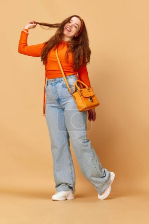 Cheerful student. Portrait of young, pretty girl wearing bright clothes, jeans touching hair with hand and smiling on ginger background. Concept of fashion, beauty, lifestyle, human emotions, ad