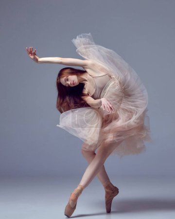 Weightless easy jump. Sensual red haired ballerina wearing tulle dress dancing with emotions over grey studio background. Contemporary dance. Concept of classical ballet, inspiration, beauty, motion