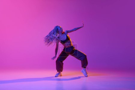 One young, attractive girl with dreadlocks dancing in street style over gradient purple neon background. Concept of contemporary dance style, inspiration, movement, motion, hobby, fashion, art, ad