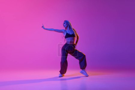 Hip-hop. Porträt mit einem jungen Mädchen, einer inspirierten Tänzerin mit Zöpfen, die mit den Händen über dem violetten Hintergrund im Neonlicht tanzt. Zeitgenössischer Tanzstil, Bewegung, Kunst, Bewegung, Werbekonzept