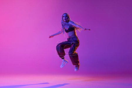 Schweben. Eine Tänzerin, eine Frau in lässiger Kleidung, springt in Bewegung über den lila Neonhintergrund. Konzept des zeitgenössischen Tanzstils, Inspiration, Bewegung, Bewegung, Hobby, Mode, Kunst