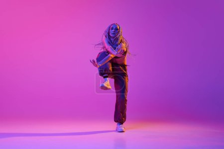 Break dance street style. One gorgeous girl, young modern dancer doing hip-hop tricks over purple studio background in neon light. Concept of contemporary style, motion, art, movement, inspiration, ad