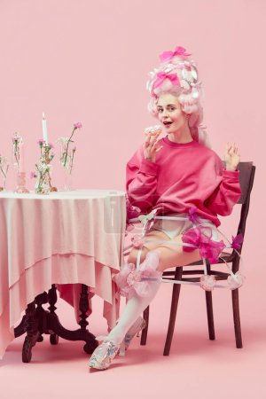 Photo for Yummy lunch. Portrait gorgeous princess, queen wearing big pink wig holding donut with exciting face on pink studio background. Comparison of eras, modernity and renaissance, beauty, food, diet, ad - Royalty Free Image