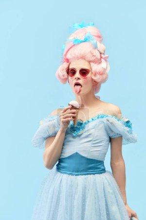 Photo for Young princess, queen wearing sunglasses eating ice cream and looking at camera over blue studio background. Concept of comparison of eras, modernity and renaissance, baroque style, beauty, fashion - Royalty Free Image