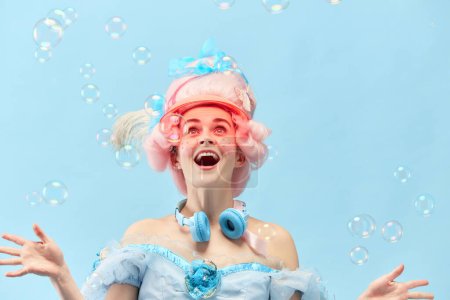 Retrato con la princesa, reina vistiendo vestido jugando con burbujas de jabón y sonriendo sobre fondo de estudio azul. Concepto de comparación de épocas, modernidad, estilo barroco, belleza, moda, emoción humana